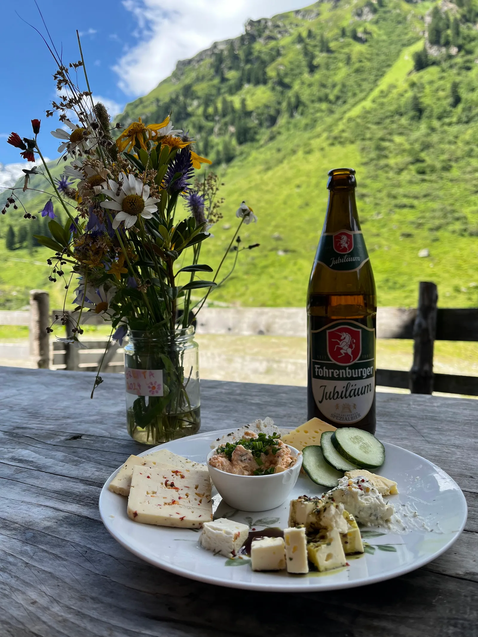 Teller mit Sura Kees, Aufstrich und Gurkenscheiben, daneben ein Strauß Wiesenblumen vor der Bergwiese